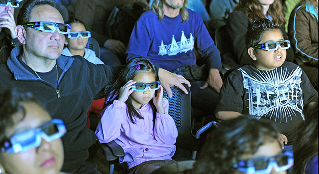 An audience wears 3-D glasses while in a darkened theater