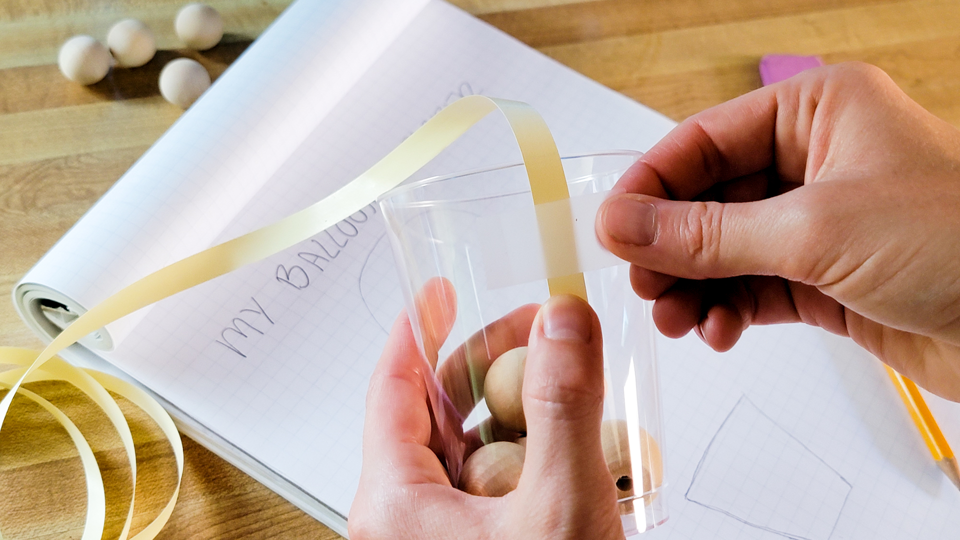 A person tapes ribbon to a cup filled with wooden beads.