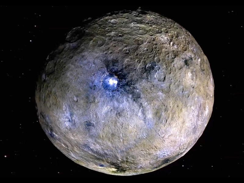 A rocky world covered with craters and bright spots appears brown with splotches of white and purple, the most intense of which is in a large central crater.