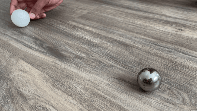 A foam ball is rolled toward a metal sphere. The foam ball shoots off in a diagonal direction after colliding with the metal sphere while the metal sphere wobbles slightly.