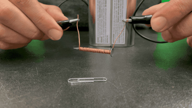 A person hovers the wire-wrapped nail portion of their electromagnet over a paperclip and picks it up using the electromagnet.