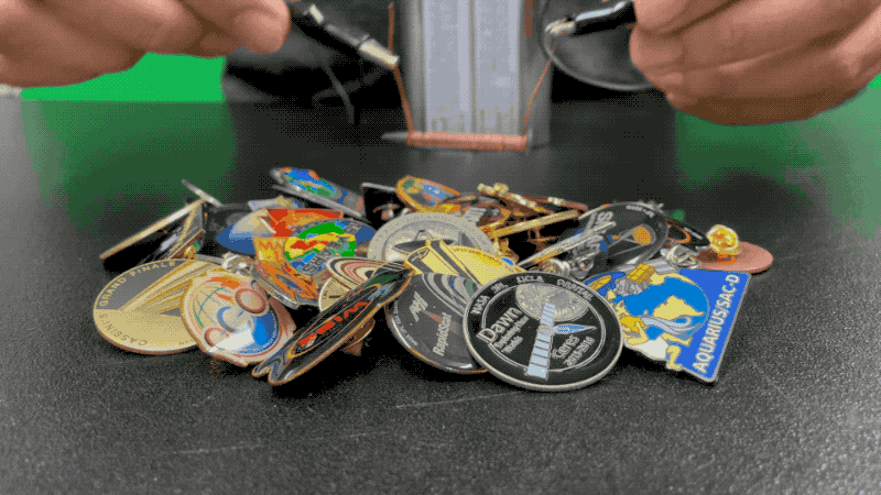 A person hovers the wire-wrapped nail portion of their electromagnet over a pile of enamel pins and picks up several of them using the electromagnet.