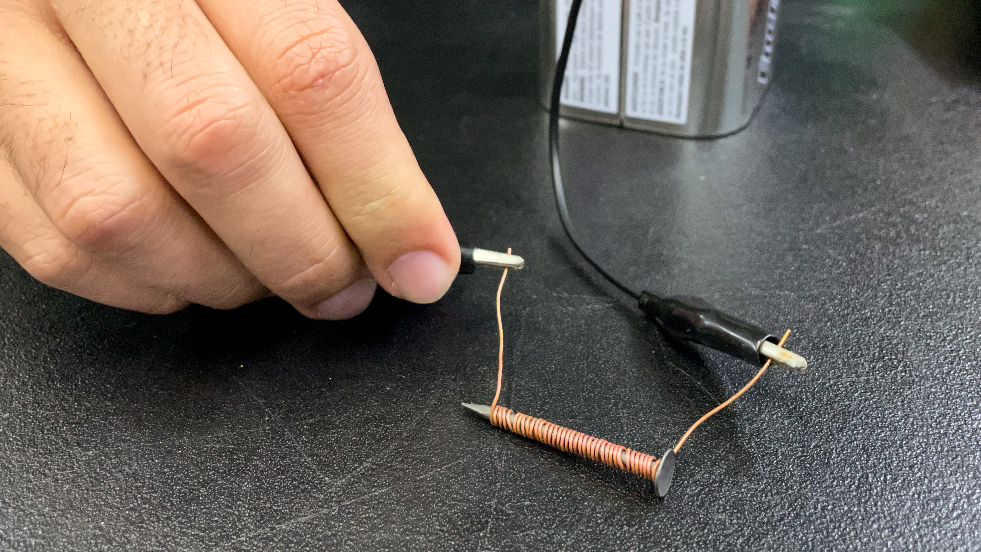 A person connects their wire-wrapped nail to a large battery using alligator clips.
