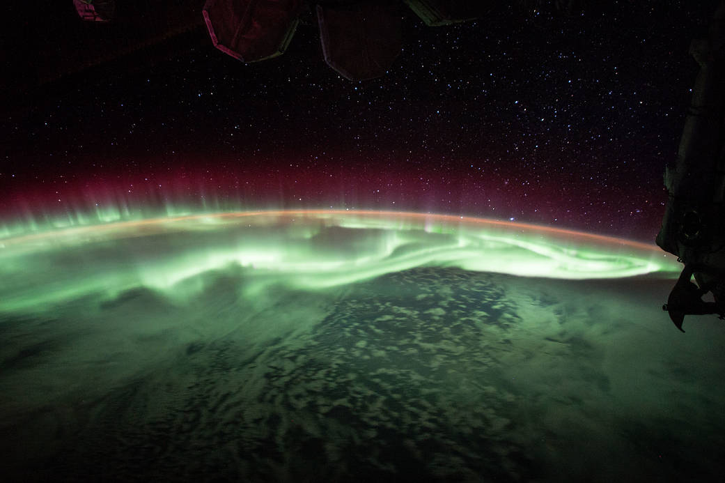 A swirling fabric of glowing neon green, orange, and pink extends above Earth's limb. A partial silhouette of the ISS frames the right corner of the image.