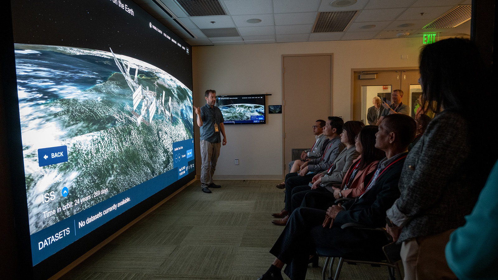 A man standing in front of an audience points to a large screen showing a simulated view of the ISS orbiting Earth.