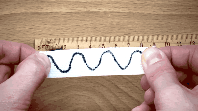 Animation of a person stretching out a piece of elastic with a wave pattern drawn on it. As the elastic gets stretched, the wavelength gets longer. 