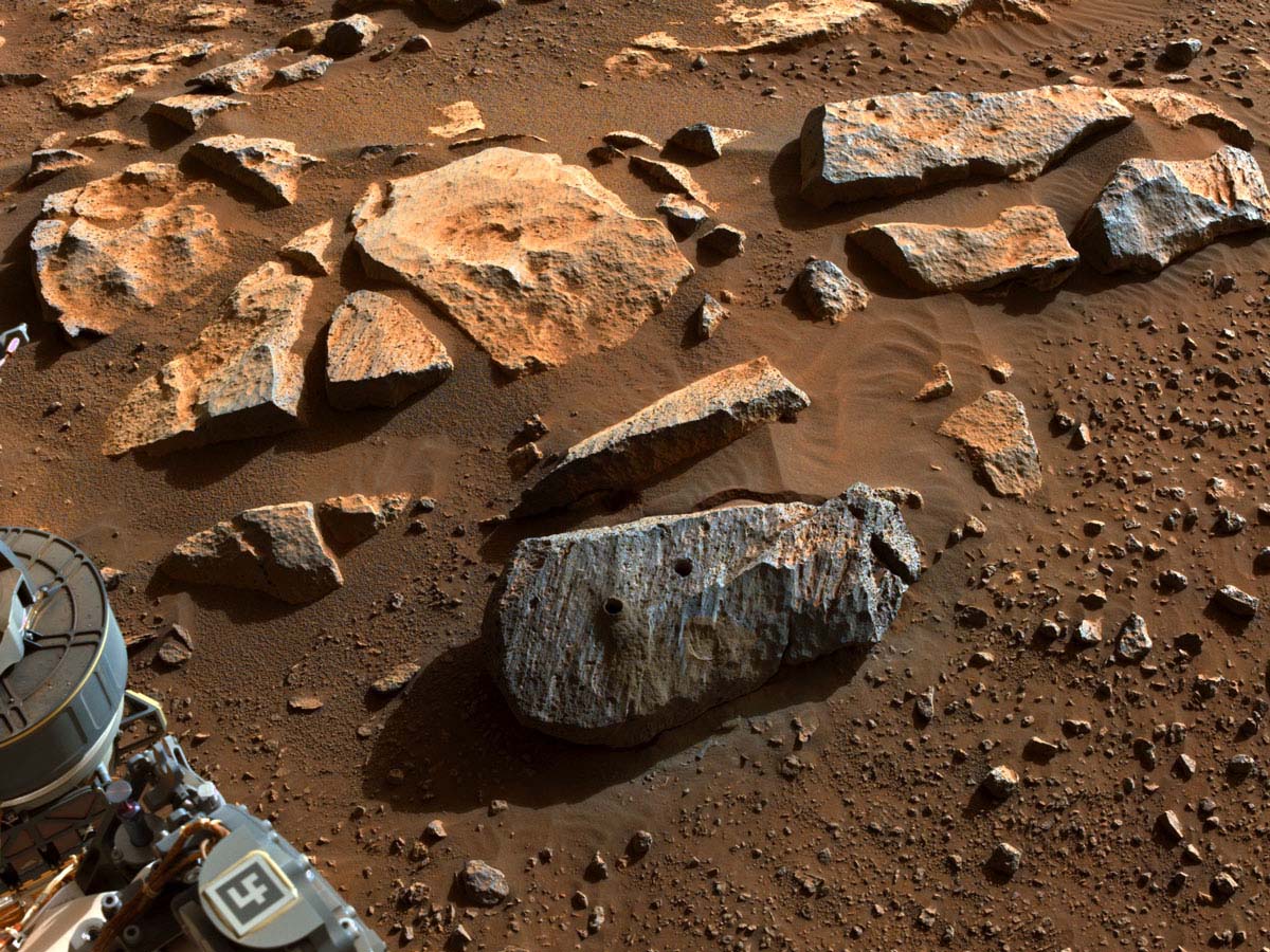 A rectangular rock with diagonal striations across its surface and two holes drilled into it is shown among reddish sand and rocks on Mars.