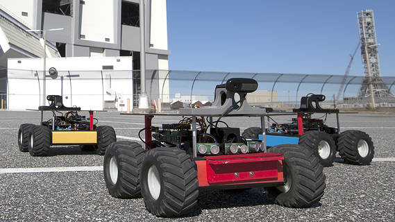 A set of small four-wheeled robots is shown on an asphalt parking lot.