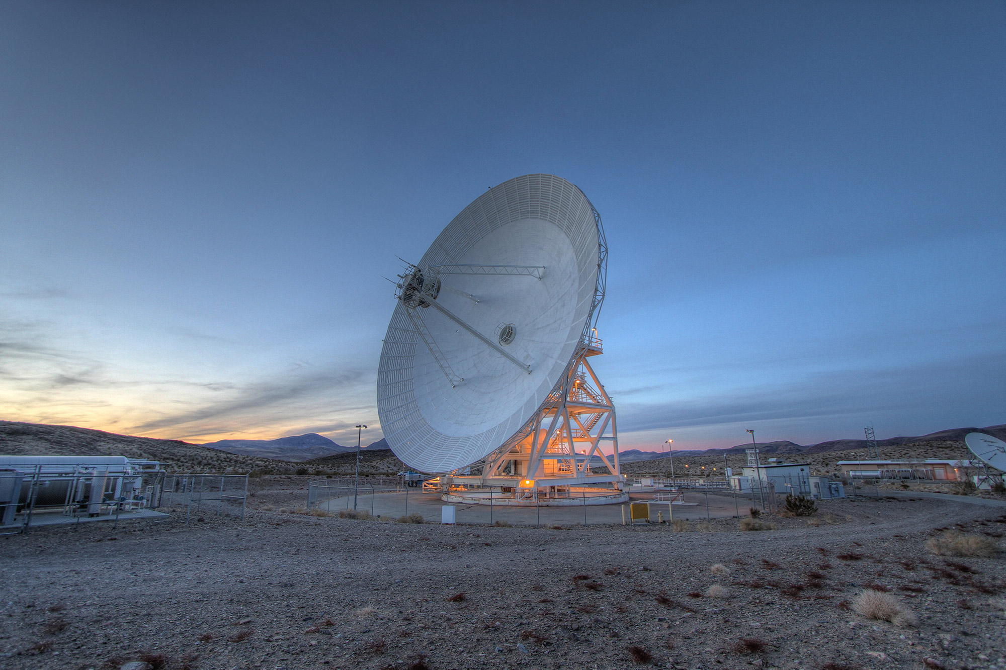 A large parabolic dish with its antenna suspended above the center by three metal supports, like legs on a stool, faces the 9 o'clock position in the image. The dish is surrounded by a desert landscape and pictured during sunset.