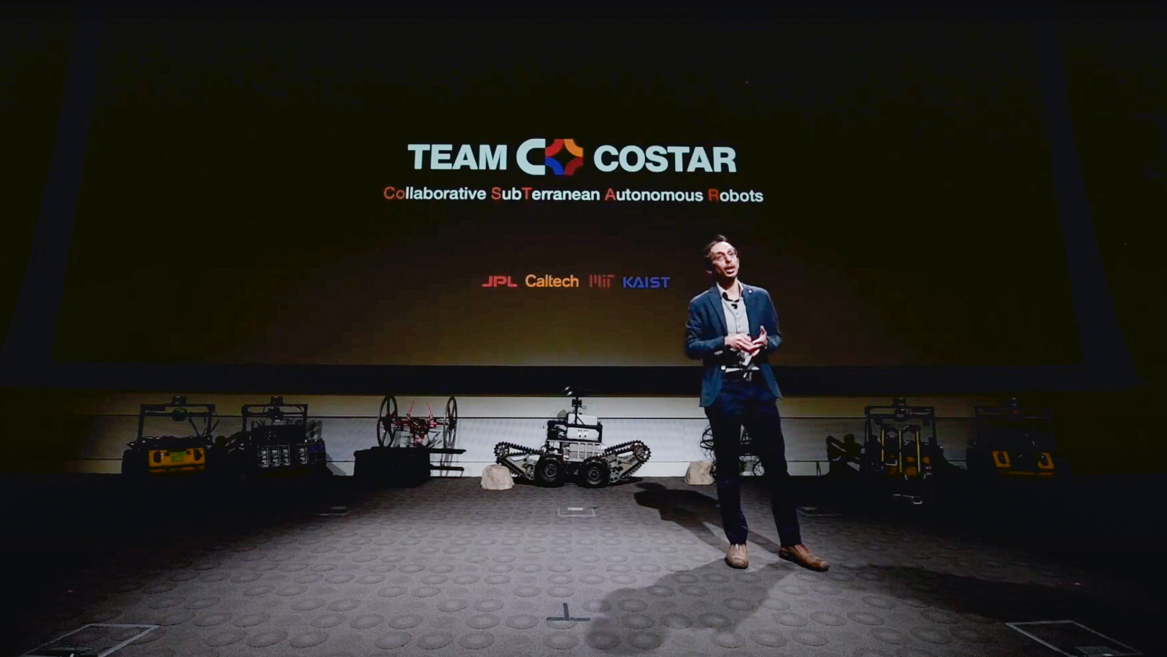 Agha stands in front of a large projector screen with robots of various shapes and sizes lined up against the wall behind him.