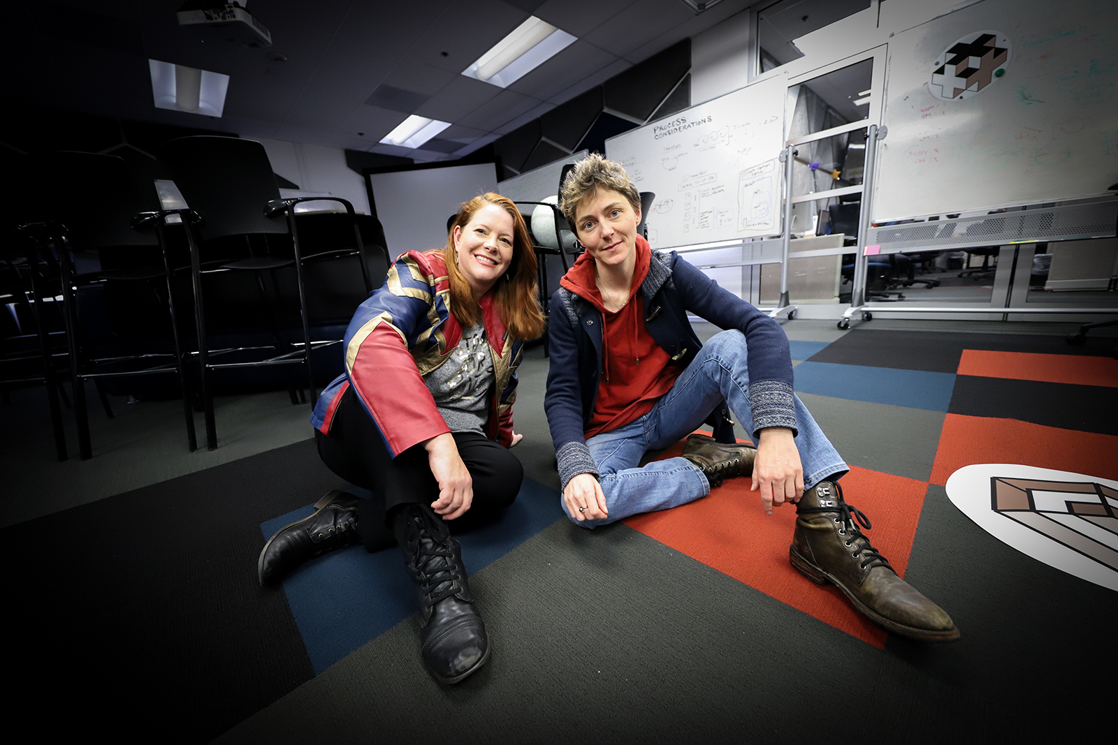 Blackwood and Holloway sit on a blue and black checkered floor with whiteboards behind them detailing process flows.