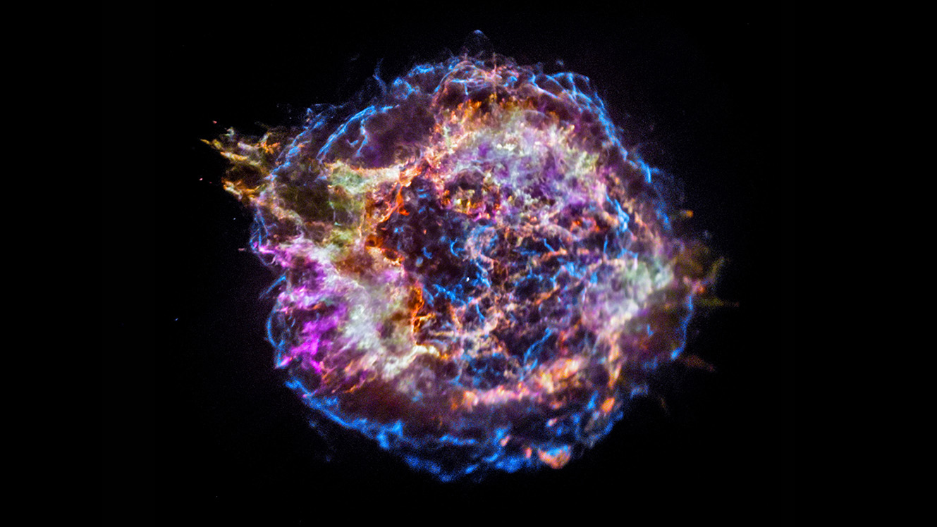 Een blauwe halo van kronkelende lijnen omringt een explosie van kleuren die zich vanuit het centrum van de supernova uitstrekken.  Het dichtst bij het centrum is een cirkelvormige oranje spetter omringd door groen en geel en tenslotte een wazig paars.