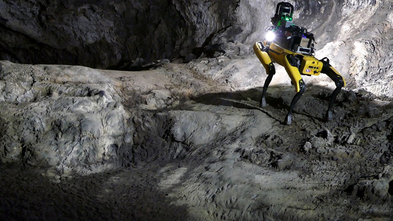 A robot shaped like a dog and carrying various tools on its back shines a light into a darkened cave.