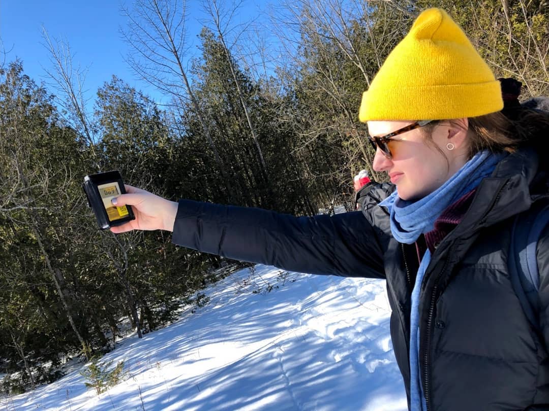 Brittney Copper stands in the snow surrounded by pine trees and holds out a device to measure the flux of solar radiation