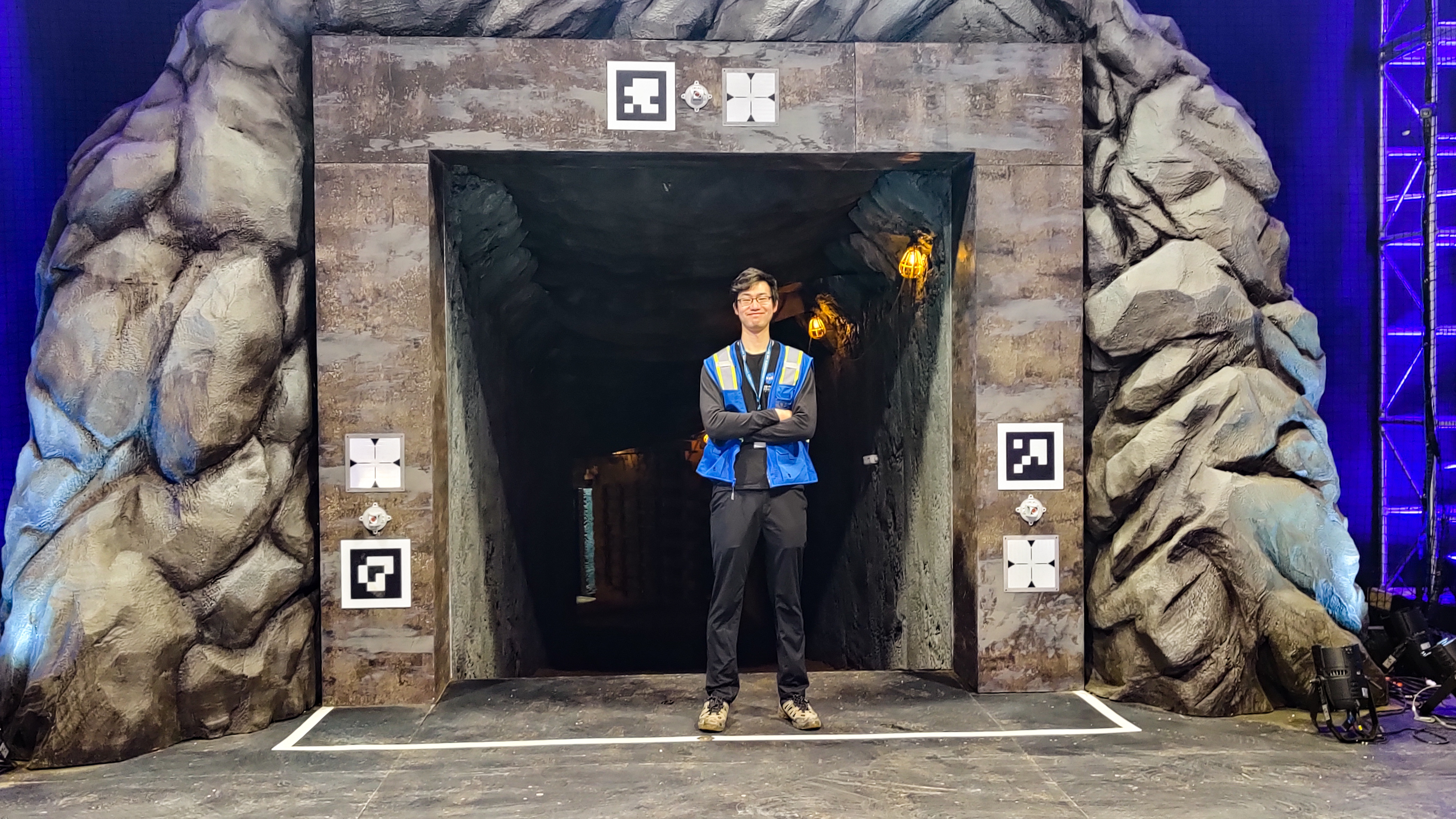 Fan stands with his arms crossed in front of a fake rock wall and spotlights framing a rocky tunnel.