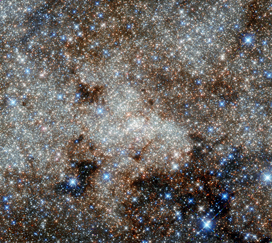 A dense field of stars like grains of sand is surrounded by wispy clouds of glowing gas and dust.