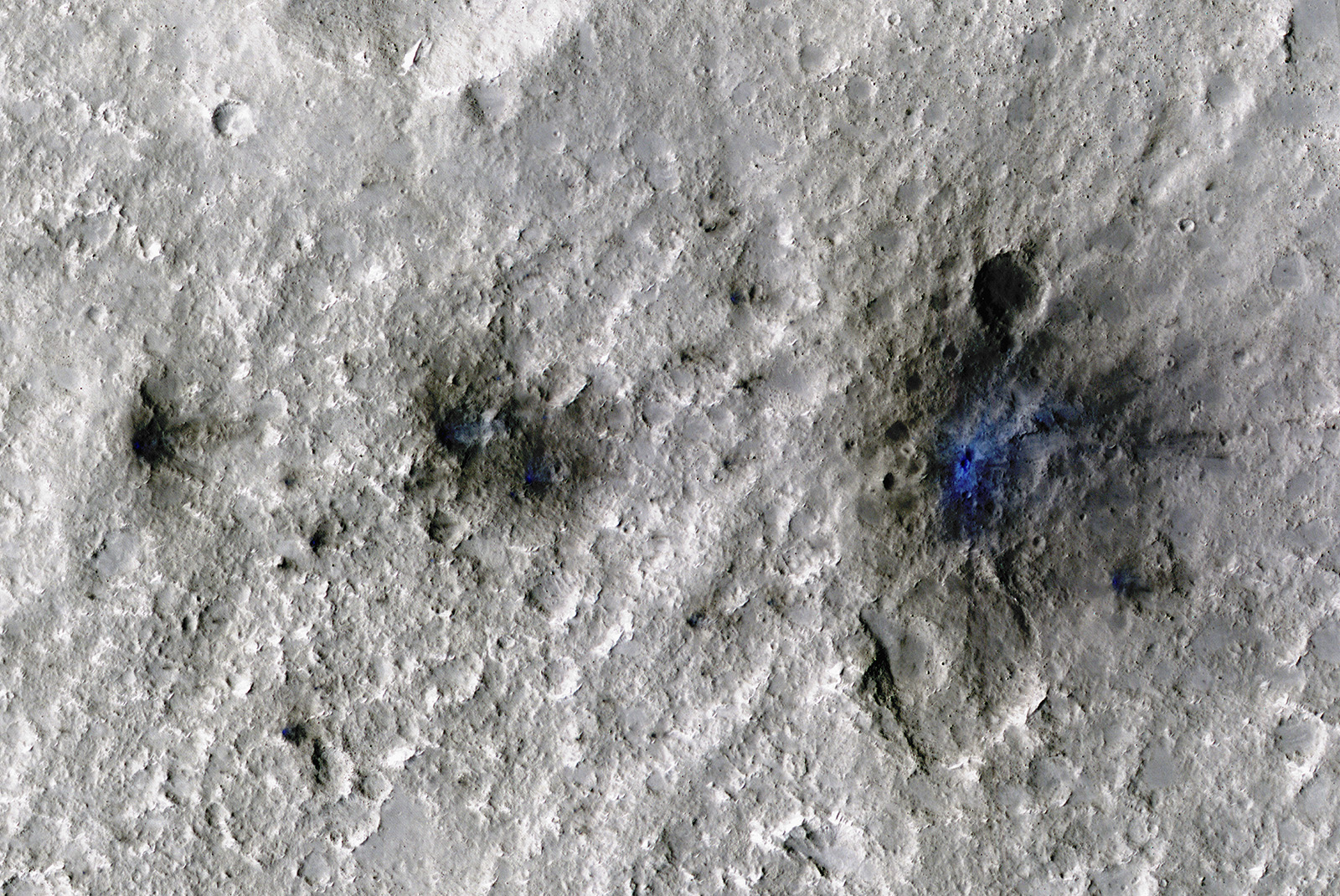 A direct overhead view of a light-gray-colored cratered surface is interrupted by three black splotches of increasing size from left to right. At the center of each dark scar is a royal blue splotch. The surface around the blue center looks as if it's been sprayed with a dark material that extends farther on the right side of each crater than on the left.
