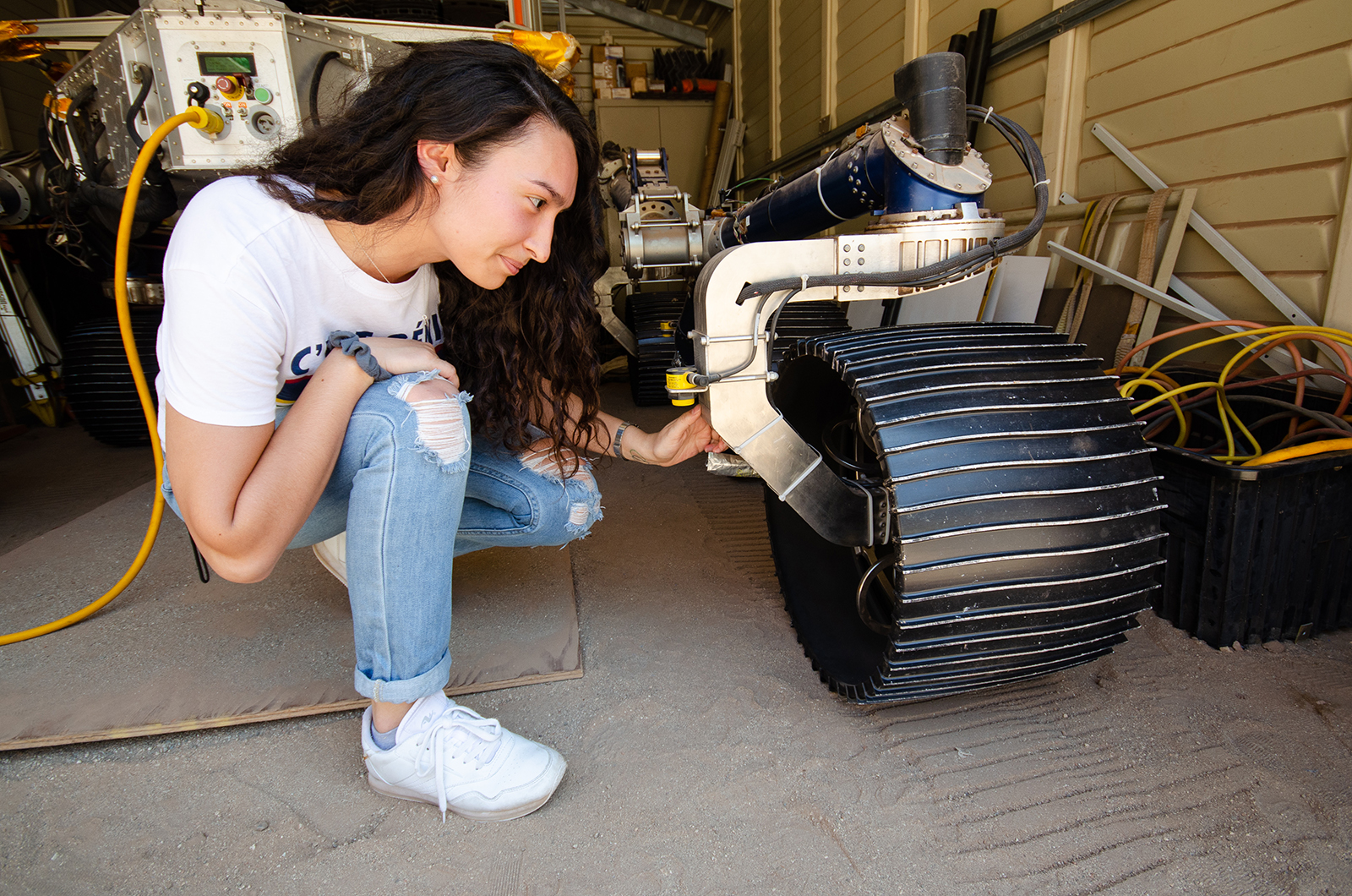 Isabel Rayas kneels down in front of the Scarecrow rover in a garage and places her hand on one of the front wheels