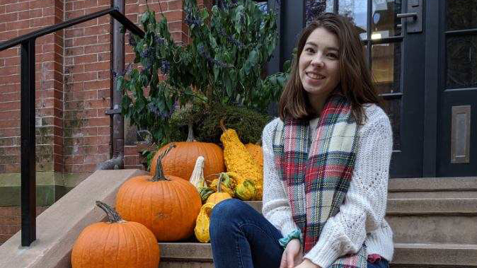 Izabella Zamora sits on steps leading up to a building with pumpkins decorating the steps to her right.