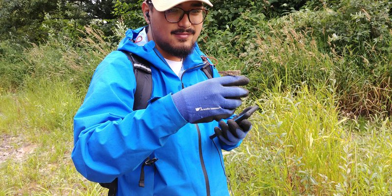 Jonathan Vellanoweth stands in a grassy field holding a phone in one hand and with a grasshopper balancing on his other hand.