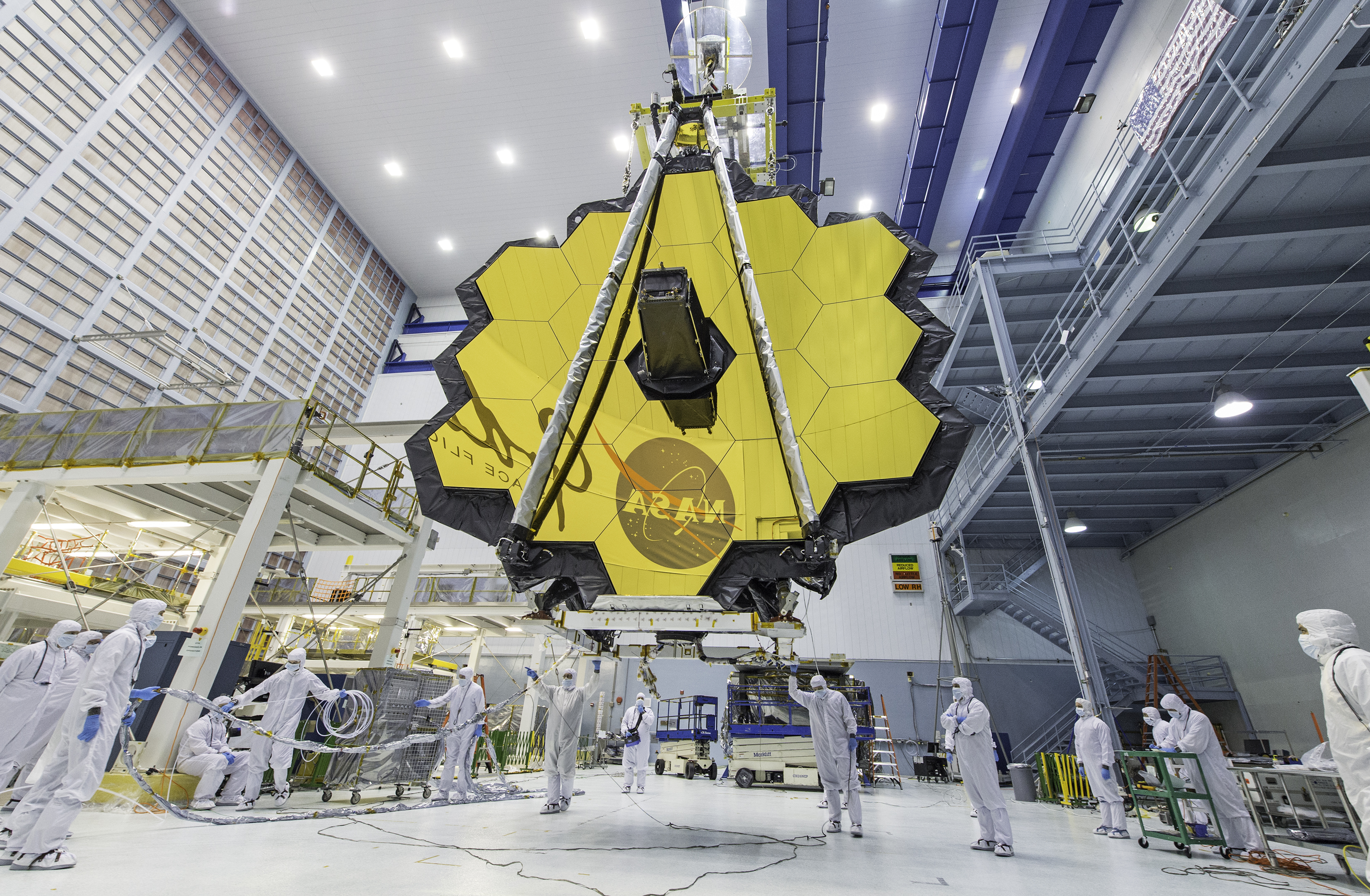 A dozen or so engineers stand in a towering open room clad in white smocks covering their entire bodies. Above them, and taking up a third of the room's space, is the reflective gold honeycomb-shaped set of mirrors for JWST. The NASA Goddard Space Center logo is reflected in the mirror.