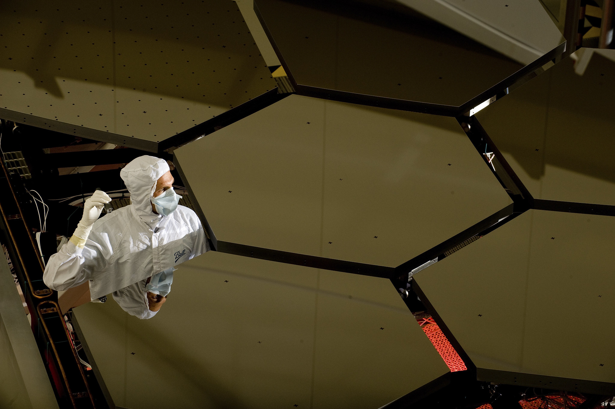 A technician in a white smock stands up in a gap between several large hexagonal mirrors forming a honeycomb shape.