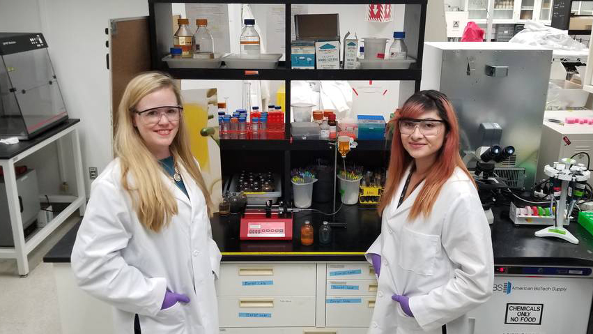 Barge and Flores pose for a photo in a lab with test tubes and scientific devices surrounding them.