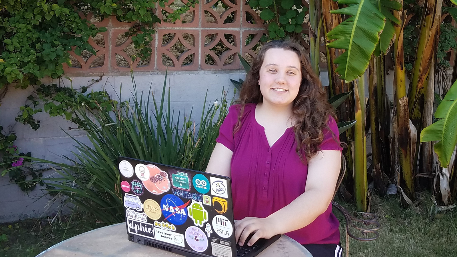 Leilani Trautman poses for a photo at an outside table. The back of her open laptop has dozens of stickers attached to it, including a NASA meatball.