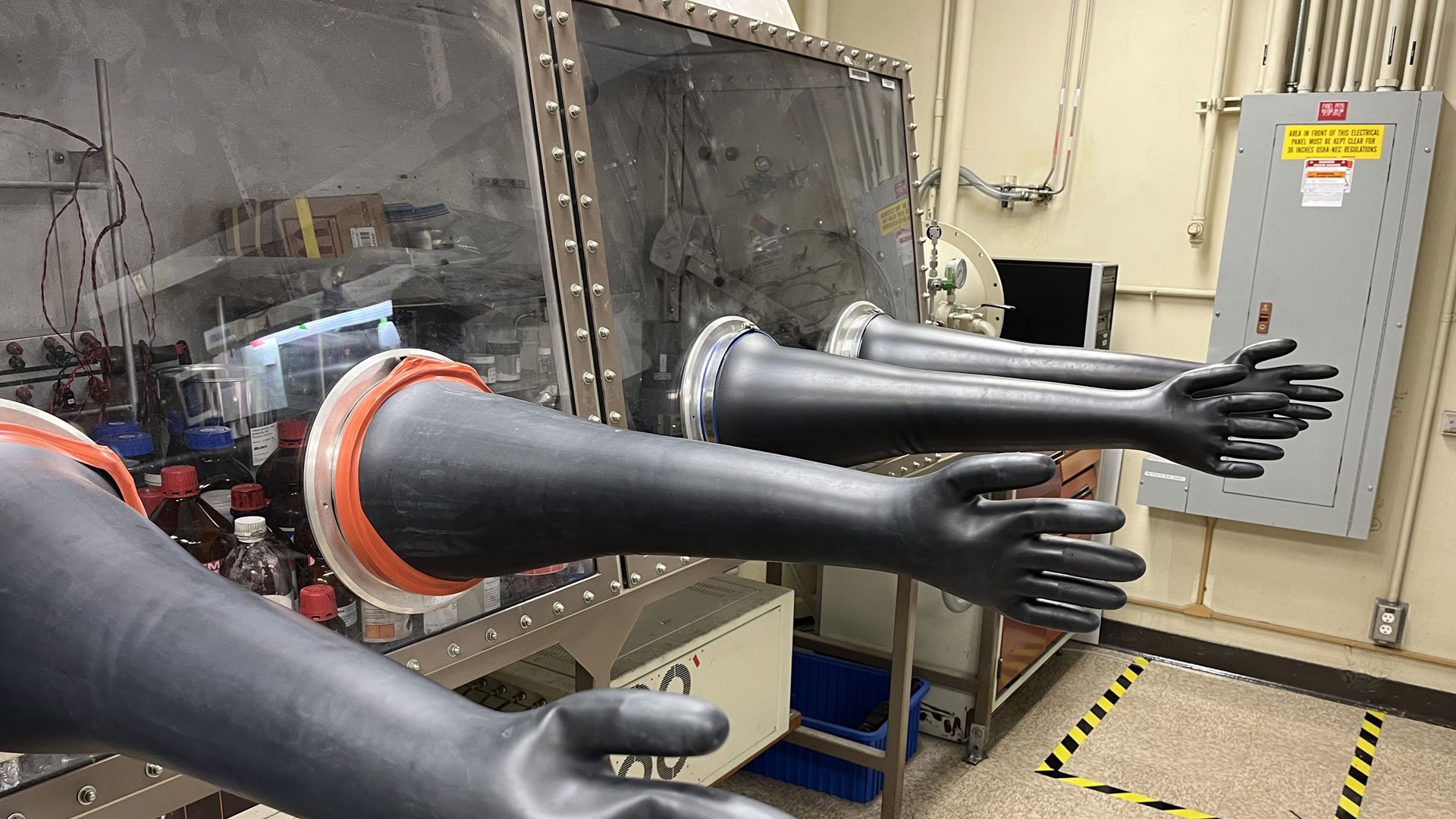 Two sets of long black gloves extend from circular openings set into the window panels of an enclosed case. Inside the case are bottles with red and blue caps, wires, and boxes.