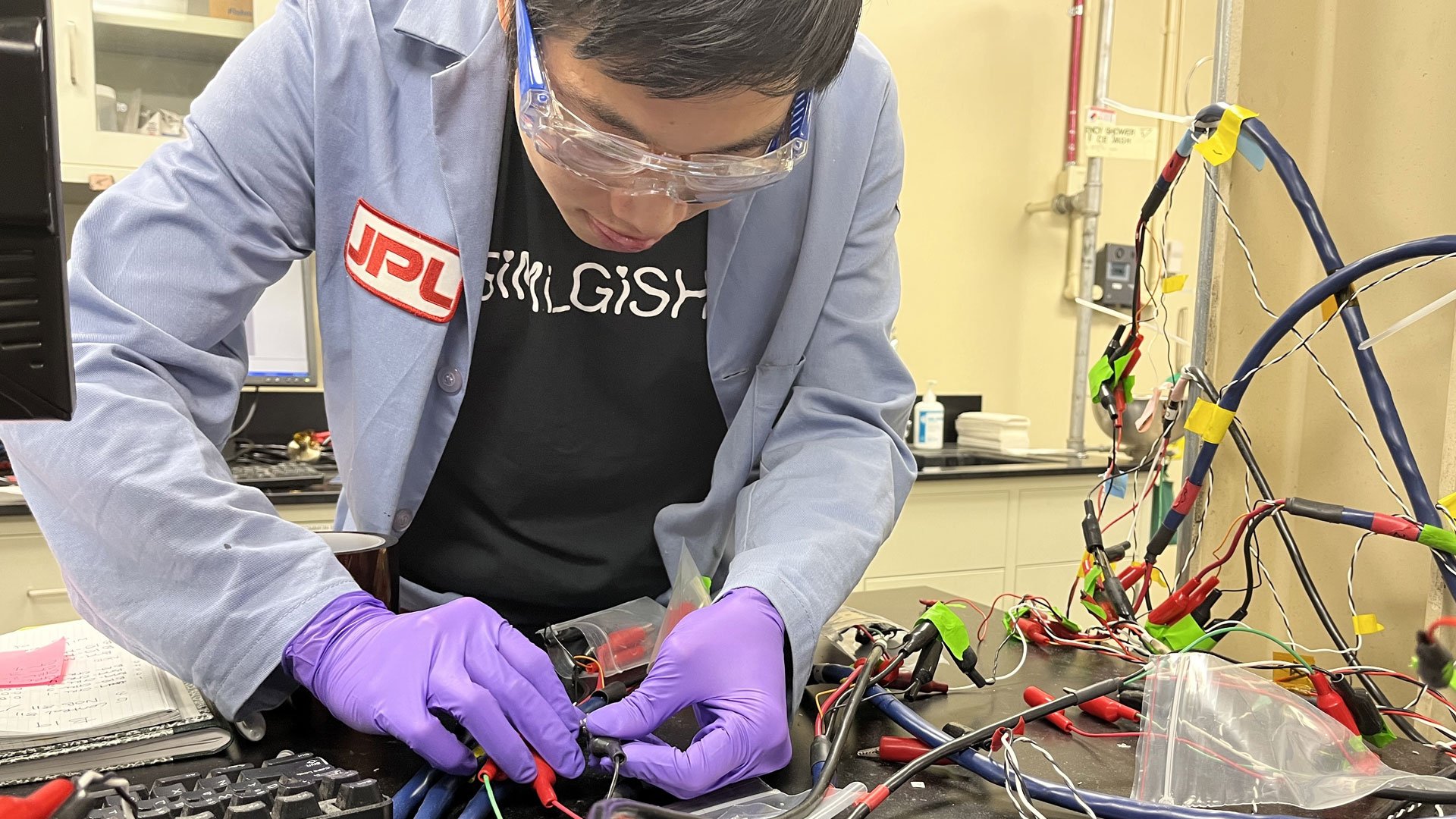 Wang wears lab goggles, a blue labcoat with a JPL patch, and purple surgical gloves and holds wires extending from a web of wires festooned with electrical tape.