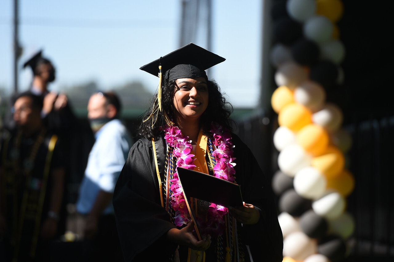 Deo wears a black cap and gown with several yellow and black cords and sashes hung around her neck along with a lei with large pink flowers.