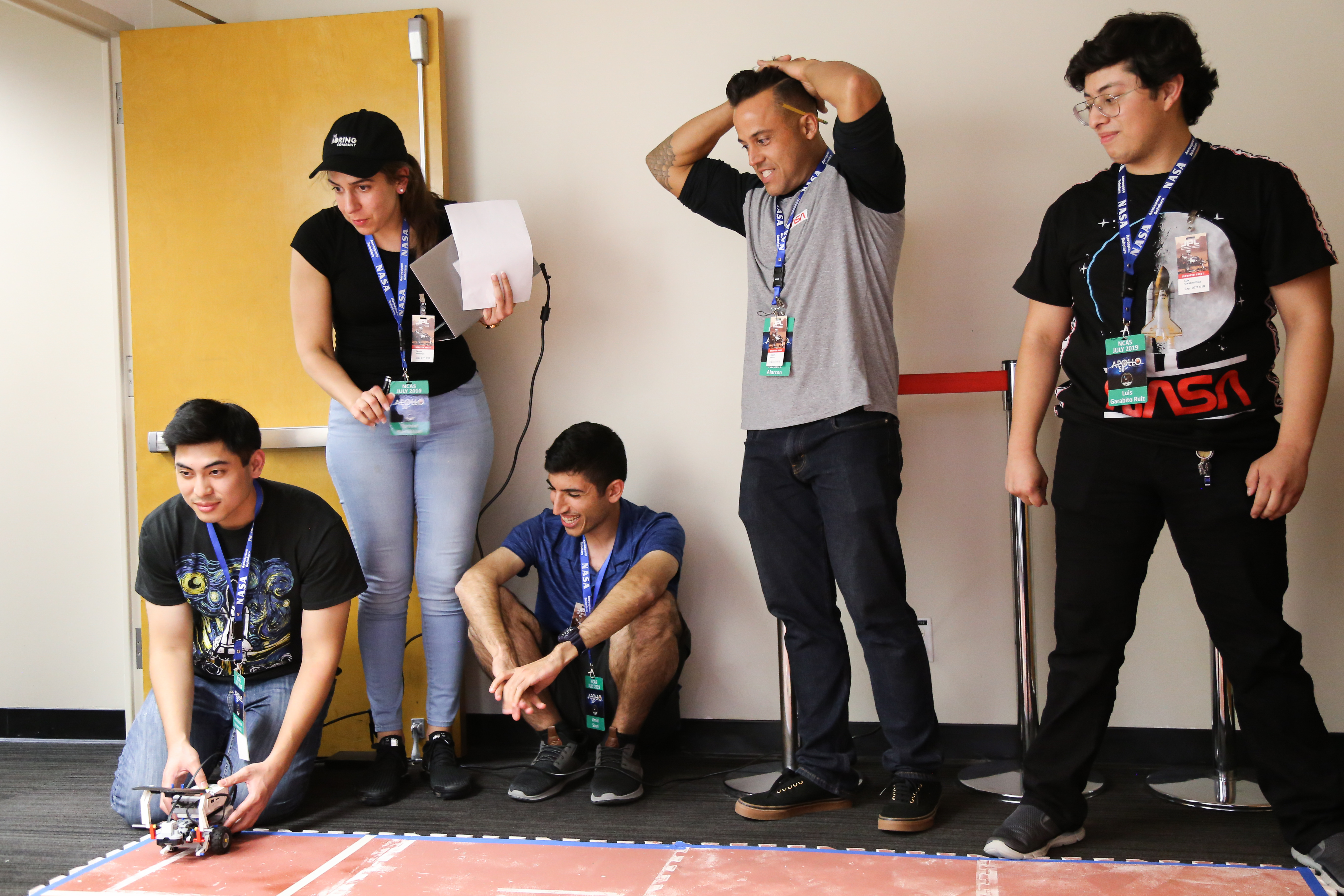 Several students stand against a wall while another sets a miniature rover on a red surface meant to simulate Martian terrain