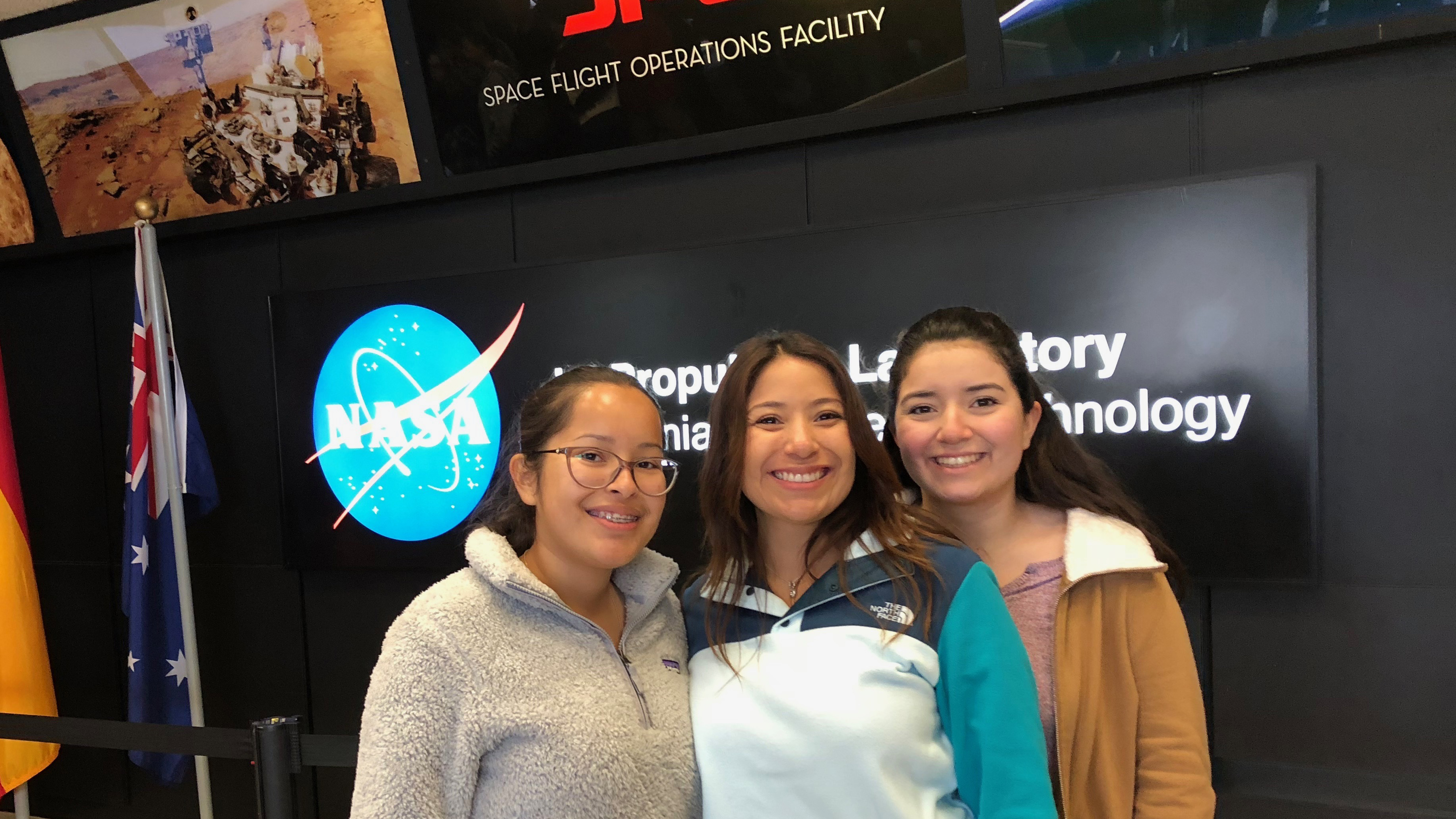 Rebecca and her mom and sister pose for a photo in the lobby of JPL's mission control with NASA/JPL logo behind them.
