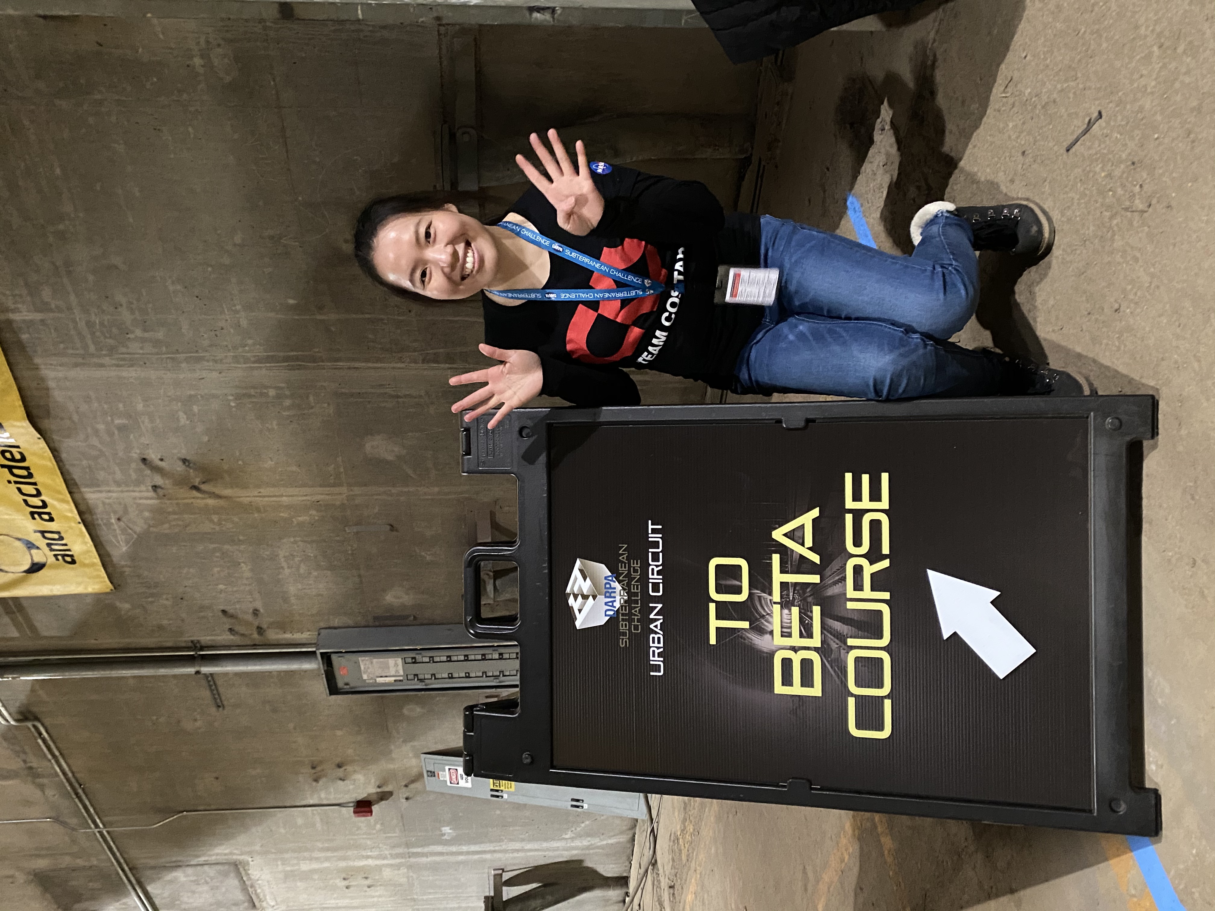 Lei wears a Team CoSTAR shirt and crouches in front of sign that reads DARPA Subterranean Challenge Urban Circuit - To Beta Course.