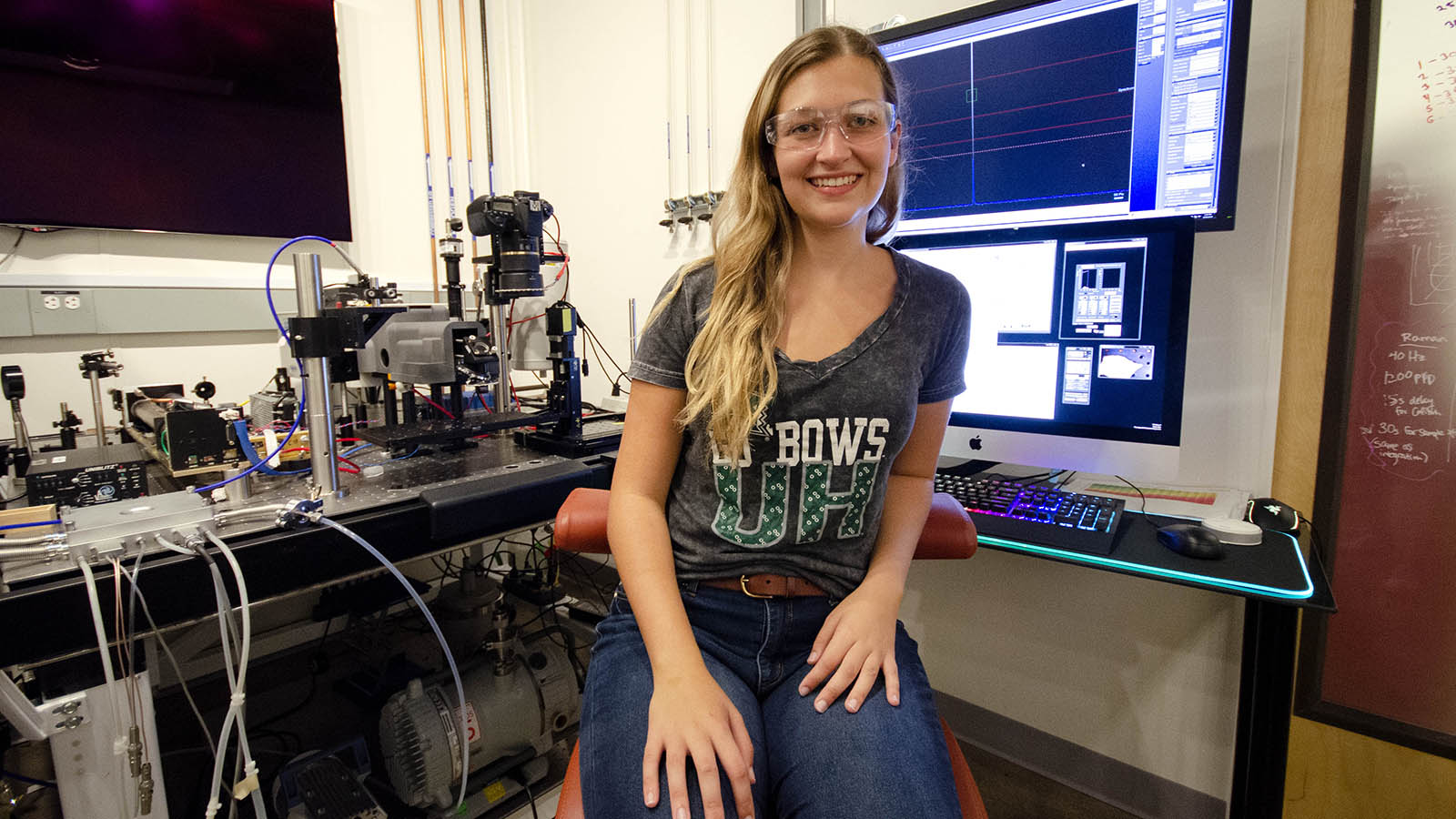 Ireland wears lab goggles and a University of Hawaii T-shirt. She sits in front of several screens and the lab version of SHERLOC.