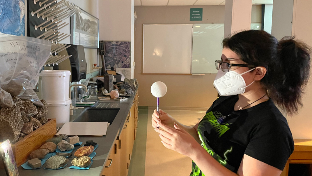 A woman wearing a mask and glasses on the right hand side looks to the left while holding a foam ball on a stick representing the moon. A light source on the left representing the Sun shines, casting a shadow on the woman's face.