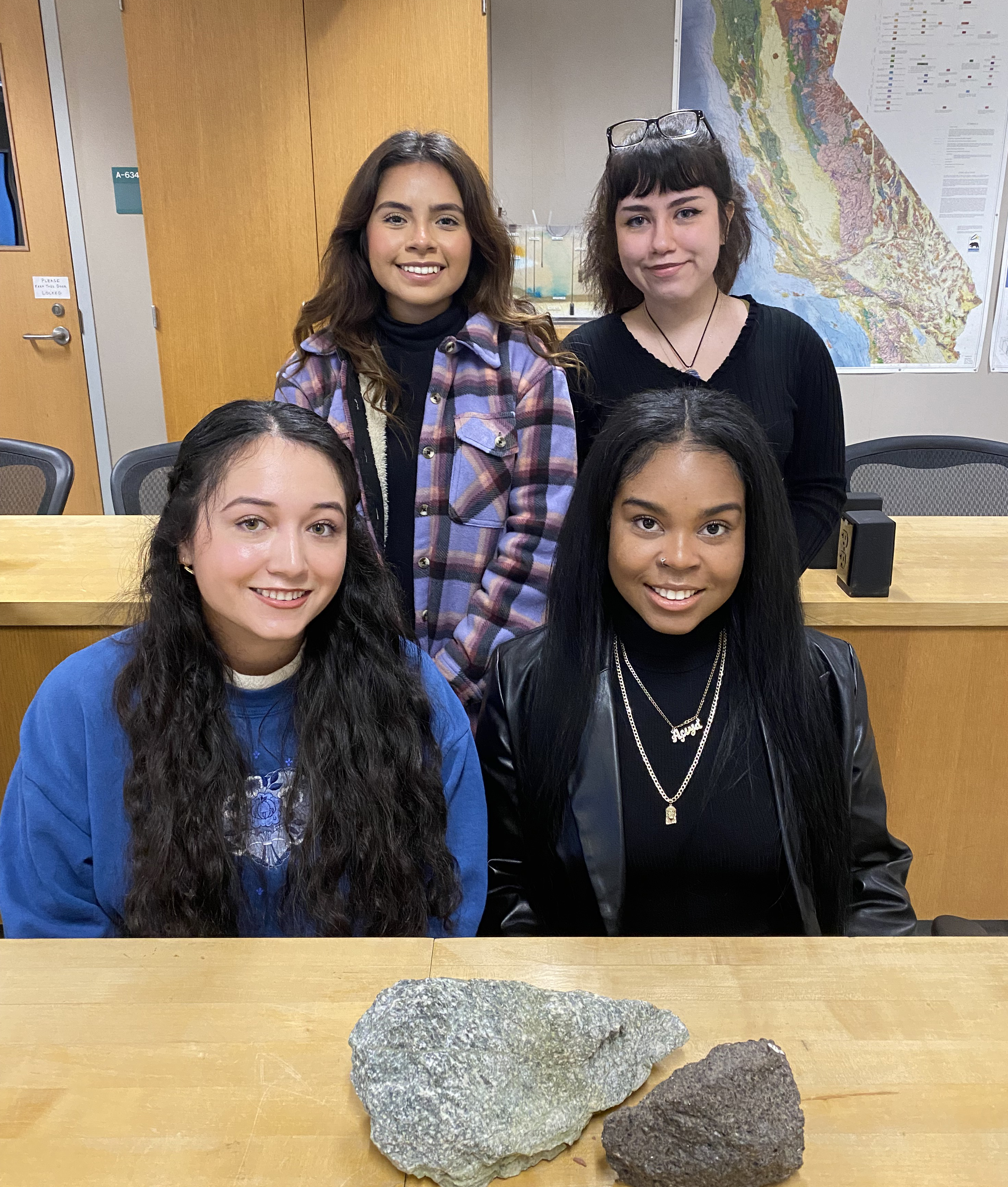 Four woman face the camera, arranged two by two, in a geology classroom. Two rocks sit on a table in front of them.