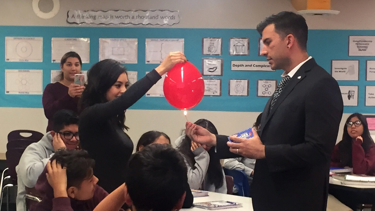 Students look on, some holding their ears, as Ms. Wisdom holds a large red balloon while NASA/JPL Education Specialist Brandon Rodriguez lights a match underneath it as part of the Global Warming Demonstration.
