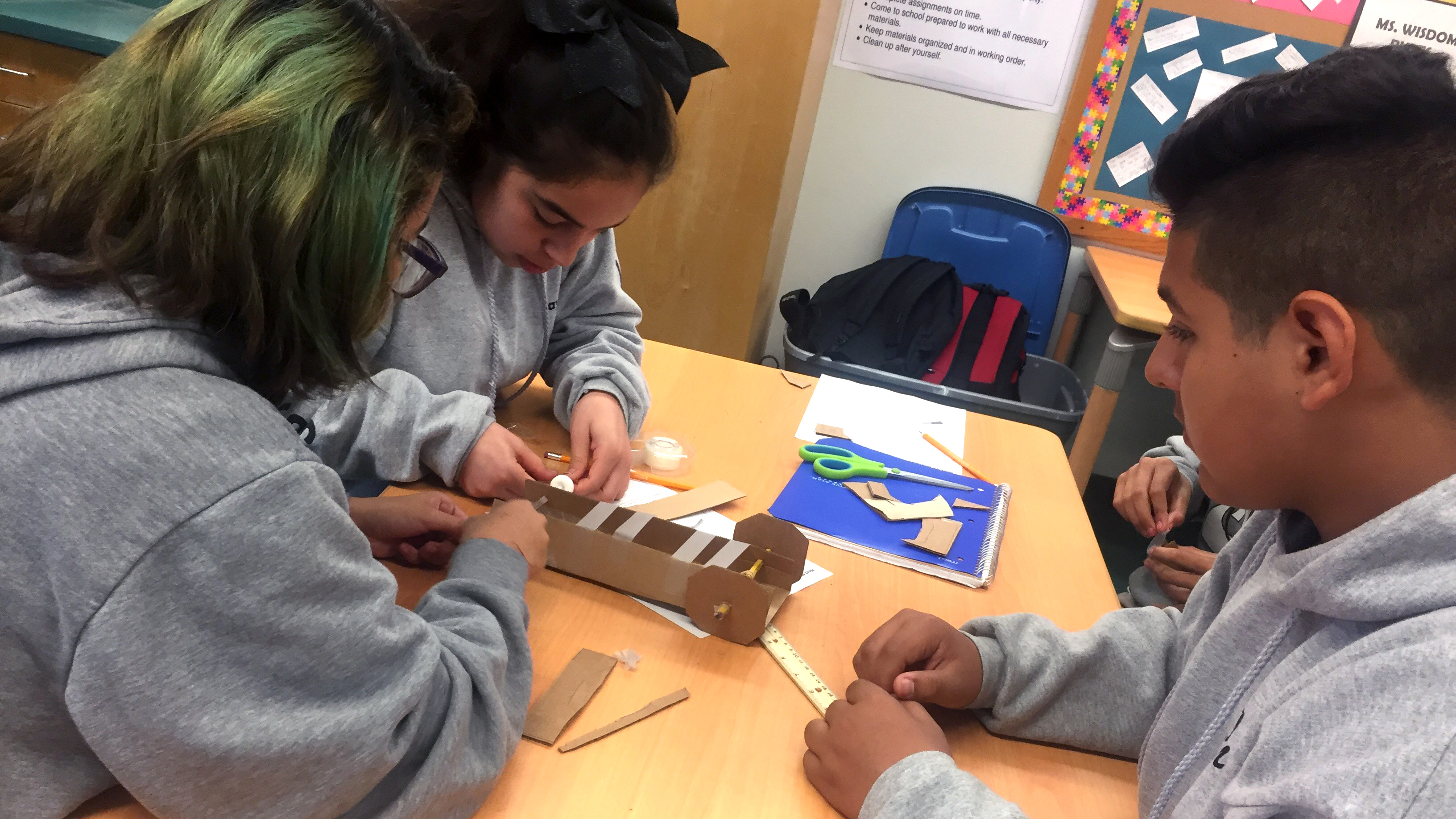 Three students in gray sweatshirts huddle around a cardboard rover, placing tape across its center.