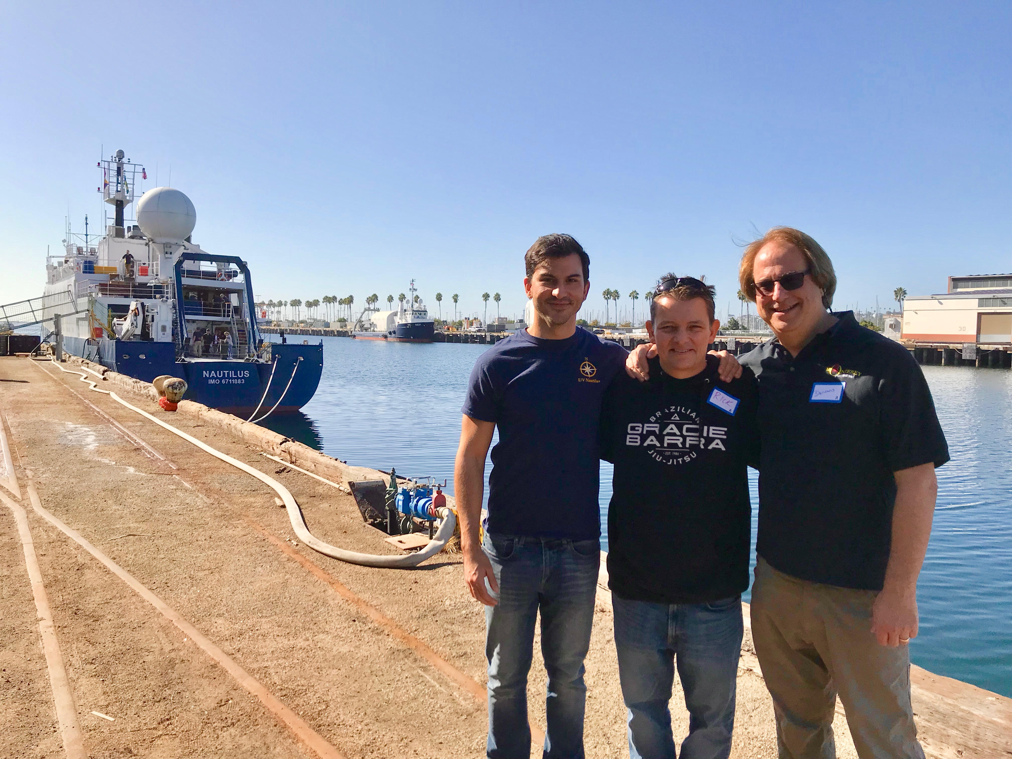 Rodriguez, Lee and Hagensmith stand on a concrete doc with a ship in the water behind them