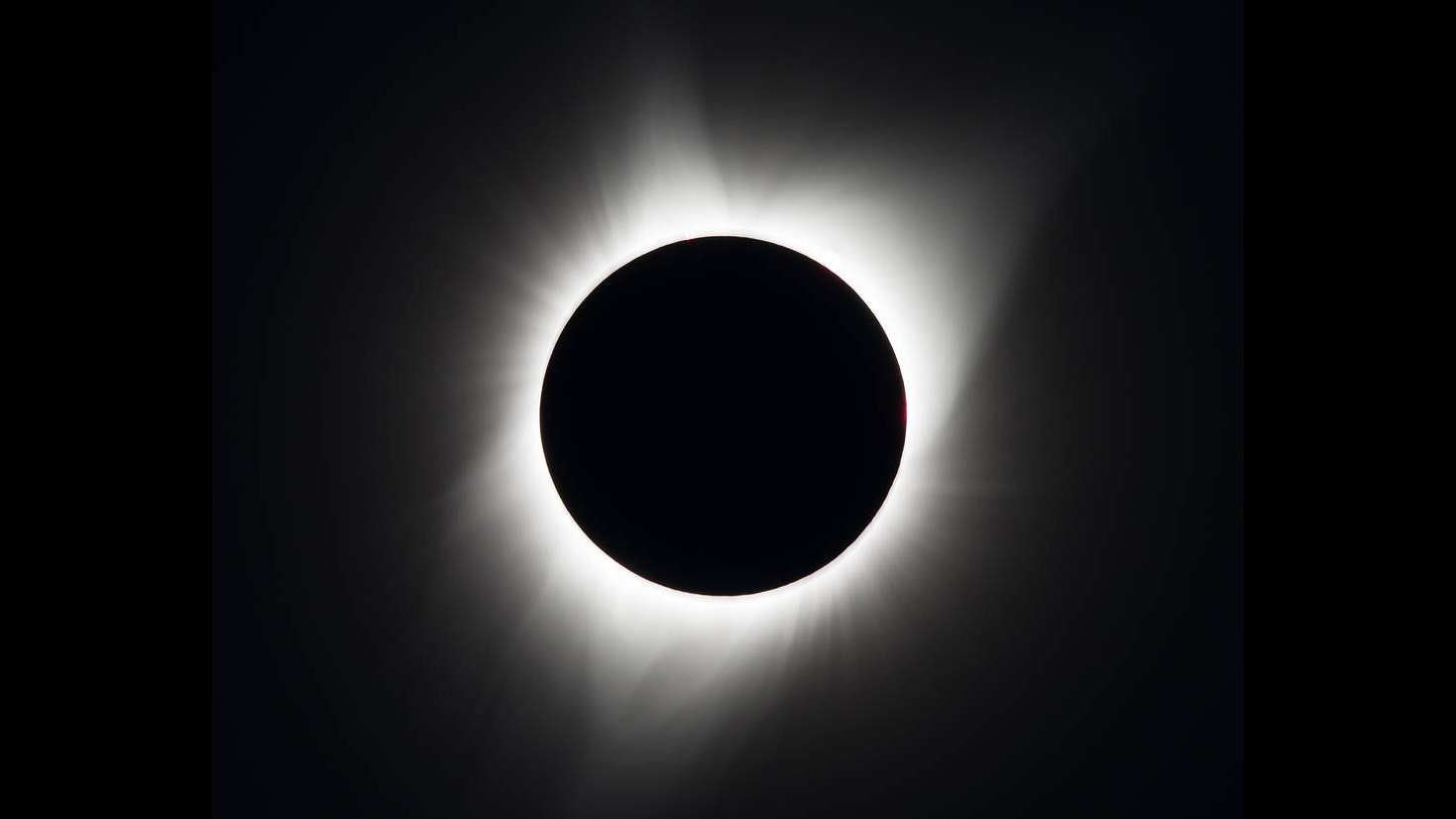 Whisps of white haze flare out around the blackened disk of the Moon, which completely covers the Sun's disk