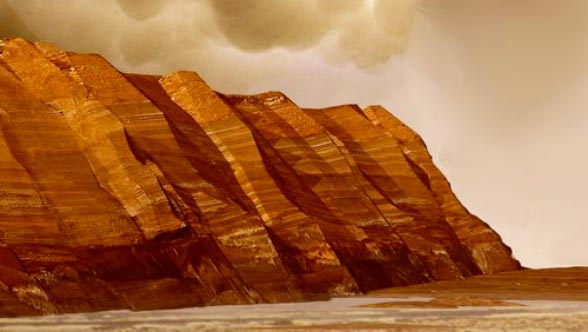 This image shows layered, red cliffs rising up from a flat, brown surface. The cliff surface slants from right to left, with the nearest cliffs on the right-hand side of the image and those farther away toward the left. Pinkish-tan clouds hover above the crests of the cliffs.